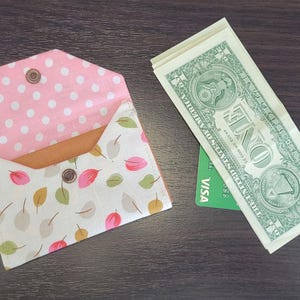 May include: A handmade fabric wallet with a pink polka dot interior and a leaf pattern exterior. The wallet is open, revealing a tan interior. Beside it are several one-dollar bills and a green credit card.