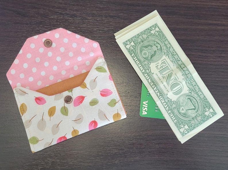 May include: A handmade fabric wallet with a pink polka dot interior and a leaf pattern exterior. The wallet is open, revealing a tan interior. Beside it are several one-dollar bills and a green credit card.