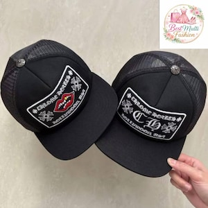 Puede incluir: Dos gorras de camionero negras con la parte trasera de malla. Cada gorra presenta un parche con el texto "Chrome Hearts Hollywood USA" y un diseño. Un parche tiene un diseño de labios rojos, el otro tiene las letras "CH".
