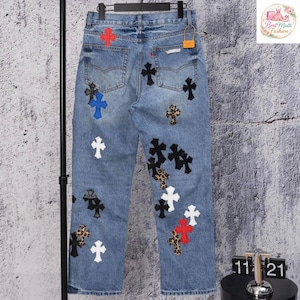 May include: Light wash denim jeans adorned with various cross appliques in black, white, red, blue, and leopard print. The jeans have a classic five-pocket design and a straight leg fit. A small orange tag is visible.