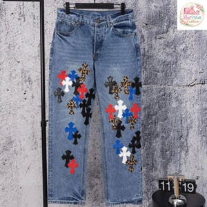 May include: Light blue denim jeans with a button fly and multiple cross appliqu&eacute;s in various colors and patterns, including black, white, red, blue, and leopard print. The jeans are displayed on a black clothing rack.