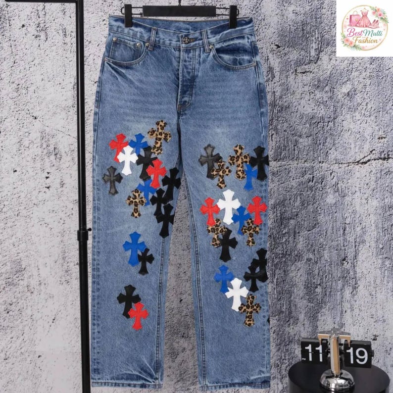 May include: Light blue denim jeans with a button fly and multiple cross appliqu&eacute;s in various colors and patterns, including black, white, red, blue, and leopard print. The jeans are displayed on a black clothing rack.