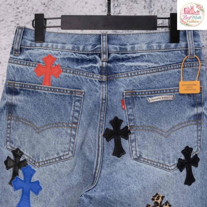 May include: Light blue denim jeans with multiple cross patches in red, black, blue, and leopard print. The jeans have a classic five-pocket design and are hanging on a black hanger.