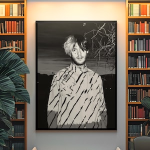 May include: Black and white portrait of a person wearing a striped shirt. The image is framed and displayed on a wall, with bookshelves on either side. The person has black and white hair.