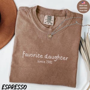May include: Espresso-colored t-shirt with the words "favorite daughter since 1981" in white script. A silver necklace with a pendant rests on the shirt. The shirt has a Comfort Colors tag.