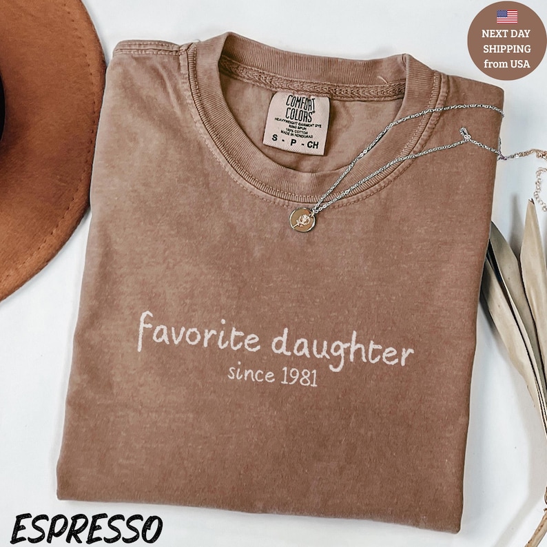 May include: Espresso-colored t-shirt with the words "favorite daughter since 1981" in white script. A silver necklace with a pendant rests on the shirt. The shirt has a Comfort Colors tag.