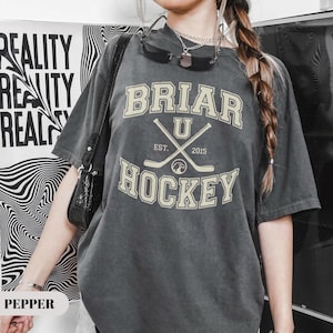 May include: Dark gray t-shirt with the text "BRIAR U HOCKEY" in a cream-colored, collegiate-style font. The design includes crossed hockey sticks and the year "2015". The shirt has a vintage, washed-out look.