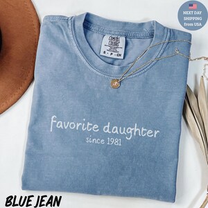 May include: Blue jean colored t-shirt with the embroidered text "favorite daughter since 1981". The shirt has a crew neck and short sleeves. A gold necklace with a pendant is draped across the shirt.