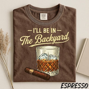 May include: Espresso-colored t-shirt with the phrase "I'll Be In The Backyard" in cream-colored script. The graphic features a whiskey glass with ice cubes and a cigar. The shirt is made of Comfort Colors material.