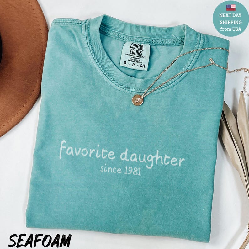 May include: Seafoam green t-shirt with the words "Favorite daughter since 1981" in white script. A gold necklace with a pendant rests on the shirt. The shirt has a Comfort Colors tag.