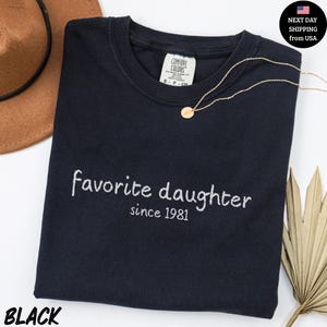 May include: Black t-shirt with the embroidered text "favorite daughter since 1981". A gold necklace with a pendant rests on the shirt. The shirt has a Comfort Colors tag. A brown hat and palm frond are in the background.