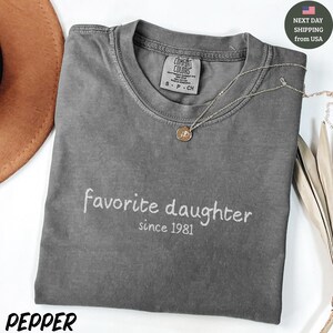May include: A gray Comfort Colors t-shirt with the words "favorite daughter since 1981" in white script. A silver necklace with a pendant rests on the shirt. The shirt is folded, and a brown hat is partially visible.
