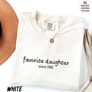 May include: White t-shirt with the text "favorite daughter since 1981" in black script. A gold necklace with a pendant rests on the shirt. The shirt is folded and placed on a white surface.