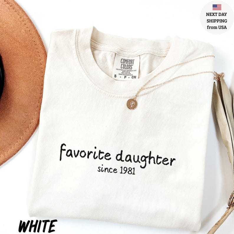 May include: White t-shirt with the text "favorite daughter since 1981" in black script. A gold necklace with a pendant rests on the shirt. The shirt is folded and placed on a white surface.
