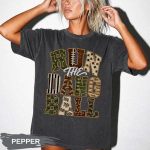 May include: A charcoal gray short-sleeve t-shirt featuring the text RUN THE DANG BALL in large block letters. The letters are filled with various football-themed patterns including field diagrams, helmets, footballs, and team text.