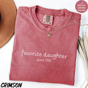 May include: Crimson-colored t-shirt with the words "Favorite Daughter since 1981" in white script. The shirt has a crew neck and a Comfort Colors tag. A silver necklace with a pendant rests on the shirt.