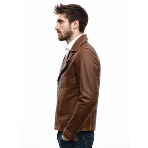 May include: A brown leather jacket with a classic collar, long sleeves, and a buttoned cuff detail. The jacket is worn over a white collared shirt and dark blue denim jeans, showcasing a smooth texture and a structured fit for the upper body.