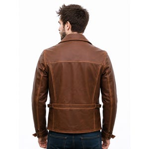 May include: A brown leather jacket featuring a wide collar, long sleeves with adjustable cuffs, and a horizontal back belt detail with side buckles. The jacket has visible seam stitching and a structured fit, worn over dark blue denim jeans.