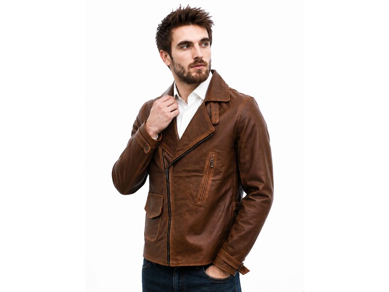May include: A brown leather jacket featuring a wide notched collar, an asymmetrical front zipper closure, and two front pockets with flaps. The jacket includes long sleeves with adjustable buckled straps at the cuffs and a white collared shirt underneath.