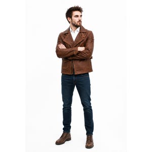 May include: A brown leather jacket with a wide collar and front zipper, worn over a white collared shirt. The outfit includes dark blue denim jeans and brown leather lace-up boots. The jacket features long sleeves and a structured fit.