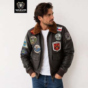 May include: A dark brown leather bomber jacket featuring a brown faux fur collar, ribbed cuffs, and multiple embroidered patches on the chest and sleeves. The jacket includes two front flap pockets and is worn over a plain white t-shirt and blue jeans.
