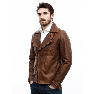 May include: A brown leather jacket featuring a wide notched collar, an asymmetrical front zipper closure, and a zippered chest pocket. The jacket includes long sleeves with buckled cuffs and a smooth finish, worn over a white collared shirt.