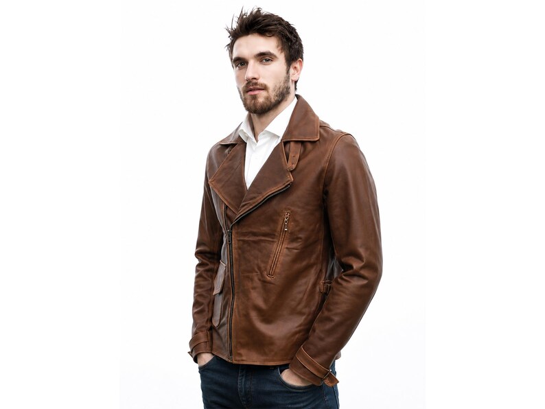 May include: A brown leather jacket featuring a wide notched collar, an asymmetrical front zipper closure, and a zippered chest pocket. The jacket includes long sleeves with buckled cuffs and a smooth finish, worn over a white collared shirt.