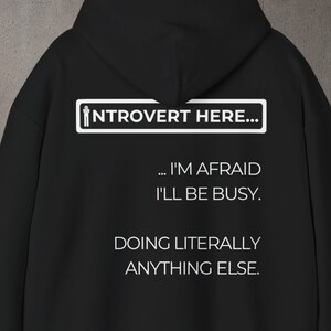 May include: Black hoodie with white text. The text reads: "INTROVERT HERE... ... I'M AFRAID I'LL BE BUSY. DOING LITERALLY ANYTHING ELSE." The hoodie has a hood and a drawstring.