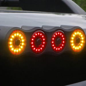 May include: Close-up of a vehicle's taillights. The lights are arranged in a row of four, with two amber lights on the ends and two red lights in the middle. Each light has a circular design with multiple LED lights.