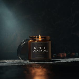 May include: A lit candle in an amber glass jar with a black lid placed to the side. The label is black with white text that reads: KNOWN & KEPT, BE STILL AND KNOW., THAT I AM GOD, PSALM 46:10, FAITH CHANGES LIVES. The candle flame glows brightly.