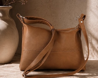 May include: A brown leather shoulder bag with a long strap. The bag has a rectangular shape with a curved top and a zipper closure. The strap is adjustable and can be worn over the shoulder or across the body. The bag is sitting on a stone surface.