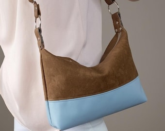 May include: A shoulder bag featuring a brown suede upper section and a smooth light blue lower panel. The bag includes a brown adjustable strap attached with silver-toned metal rings and rivets, providing a structured yet flexible design.