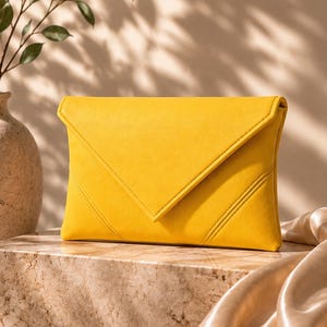 May include: A bright yellow envelope-style clutch bag featuring a triangular flap closure and two diagonal stitched lines on the lower right corner. The bag has a smooth, matte texture and a structured rectangular shape with clean, defined edges.