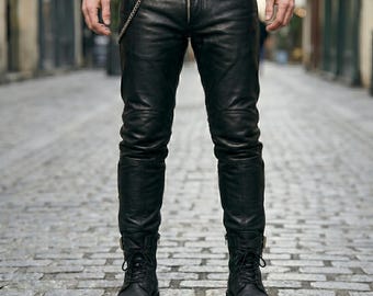 Op de afbeelding: Zwarte leren broek met ritssluiting en kettingdetail. De broek heeft versterkte kniepanelen en wordt gecombineerd met zwarte veterlaarzen. De foto is genomen op een geplaveide straat.