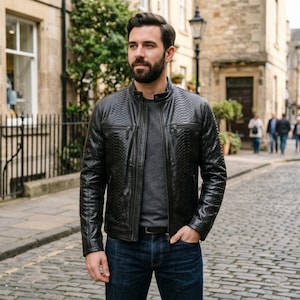 May include: A black leather jacket with a textured, embossed pattern. The jacket has a zip-up front, two chest pockets, and a stand-up collar. The person is wearing a gray shirt and blue jeans. The background is a cobblestone street.