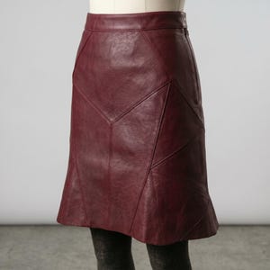 May include: A burgundy leather skirt with a geometric design. The skirt has a fitted waist and a slight flare at the hem. The panels are stitched together, creating a unique pattern. The skirt is displayed on a mannequin.