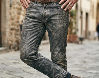 Op de afbeelding: Verweerde grijze denim jeans met een bruine leren riem en gesp. De jeans hebben een vervaagde, versleten uitstraling. De persoon draagt bruine leren laarzen. De achtergrond is een geplaveide straat.