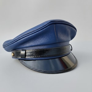 Op de afbeelding: Een marineblauwe leren hoed met een zwarte lakleren klep en band. De hoed heeft een afgeronde kroon en een kleine, goudkleurige knoop aan de zijkant. Een klassieke hoed, geschikt voor diverse gelegenheden.