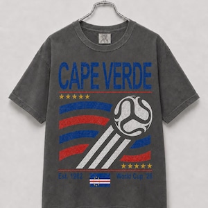 May include: A charcoal grey short-sleeve t-shirt featuring the text CAPE VERDE in blue. The graphic includes a white football, red and blue wavy stripes, ten yellow stars, the text Est. 1982 World Cup '26, and a small Cape Verde flag.