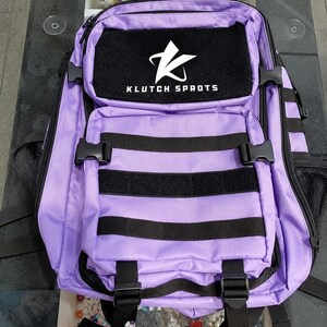 Peut inclure: Un sac à dos violet avec des sangles et des détails noirs. Le sac comporte plusieurs compartiments et un patch noir avec le texte "KLUTCH SPORTS" et un logo blanc. Il possède des sangles et des boucles réglables.