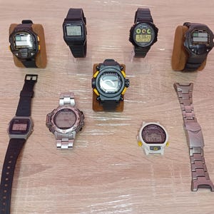 Può includere: Una collezione di orologi digitali in vari stili e colori. Gli orologi presentano dettagli neri, argentati e gialli. Alcuni orologi sono esposti su supporti marroni, altri sono piatti. L'immagine mostra una varietà di cinturini.