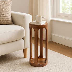 May include: A round, wooden side table with a warm brown finish. The table has a cylindrical shape with vertical cutouts and a flat top. A white mug rests on a book on top of the table. The table is next to a cream-colored sofa.