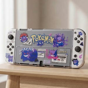 Pode incluir: Uma consola Nintendo Switch com uma capa protetora transparente com gráficos temáticos de Pokémon. A capa tem desenhos de personagens Gengar roxos e azuis, o logotipo Pokémon e texto japonês. A consola está sobre uma mesa de madeira.