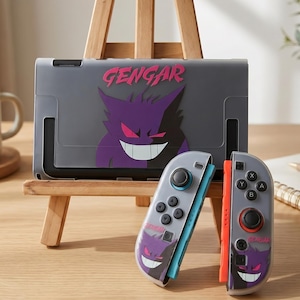 Pode incluir: Uma capa e comandos cinzentos para Nintendo Switch com um design Gengar roxo. A capa tem a palavra "Gengar" em rosa. Os comandos têm um detalhe azul claro e laranja.