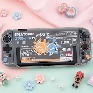 May include: A gray Splatoon-themed Nintendo Switch controller with orange and blue ink splat graphics. The controller has black buttons and a screen displaying game information. Decorative flowers, tape rolls, and figurines surround the controller.