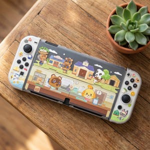 Puede incluir: Una consola Nintendo Switch con una funda protectora transparente con un diseño de dibujos animados colorido. La funda muestra varios personajes y edificios de la serie de videojuegos Animal Crossing. La consola descansa sobre una superficie de madera.