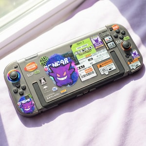 以下が含まれることがあります： グレーのNintendo Switchコンソール。ポケモンキャラクターのゲンガーのステッカーでカスタマイズされています。ステッカーには日本語のテキストと様々なデザインが含まれており、紫色のゲンガーのグラフィックが目立っています。コンソールには黒いボタンとカラフルなジョイスティックアクセントがあります。