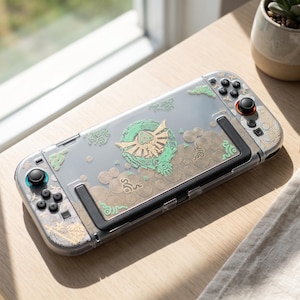 May include: A handheld gaming console featuring a clear protective case with gold and mint green graphic designs. The console includes black joysticks, buttons, and a central screen, with intricate circular and geometric patterns printed on the shell.