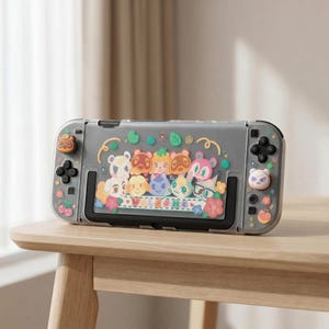 Könnte beinhalten: Eine graue Nintendo Switch-Hülle mit bunten Cartoon-Charakteren und dem Wort "WELCOME". Die Hülle hat eine transparente Vorderseite und dekorative Knöpfe. Die Hülle liegt auf einem Holztisch.