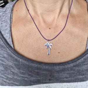 May include: A silver-colored palm tree pendant hangs from a thin, purple cord necklace. The pendant is a detailed silhouette of a palm tree. The necklace is worn against a gray shirt.